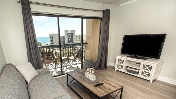 Ein Wohnzimmer mit grauem Sofa, einem Holztisch, einem Fernseher auf einem weißen Schrank und einem Balkon mit Blick auf Gebäude und das Meer.