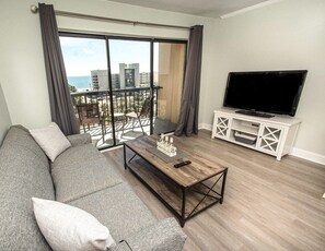 Una sala de estar con un sofá gris, una mesa de centro de madera, un televisor sobre un mueble blanco y un balcón con vistas a edificios y al mar.
