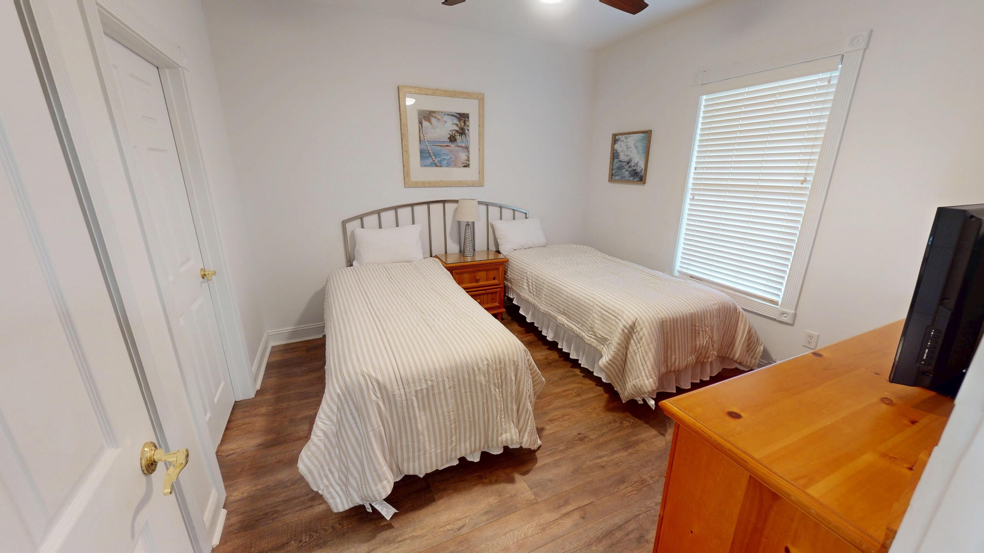 A bedroom with two beds, a wooden dresser, a ceiling fan, and a window with blinds.