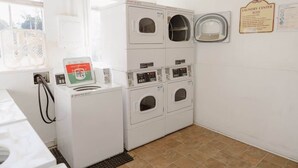 Une buanderie équipée de machines à laver et de sèche-linge superposés, d’un ventilateur et d’un panneau indiquant les règles à respecter pour l’utilisation des machines.