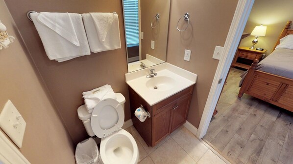 Baño con inodoro, lavabo y espejo. Se ve una cama en la habitación contigua.