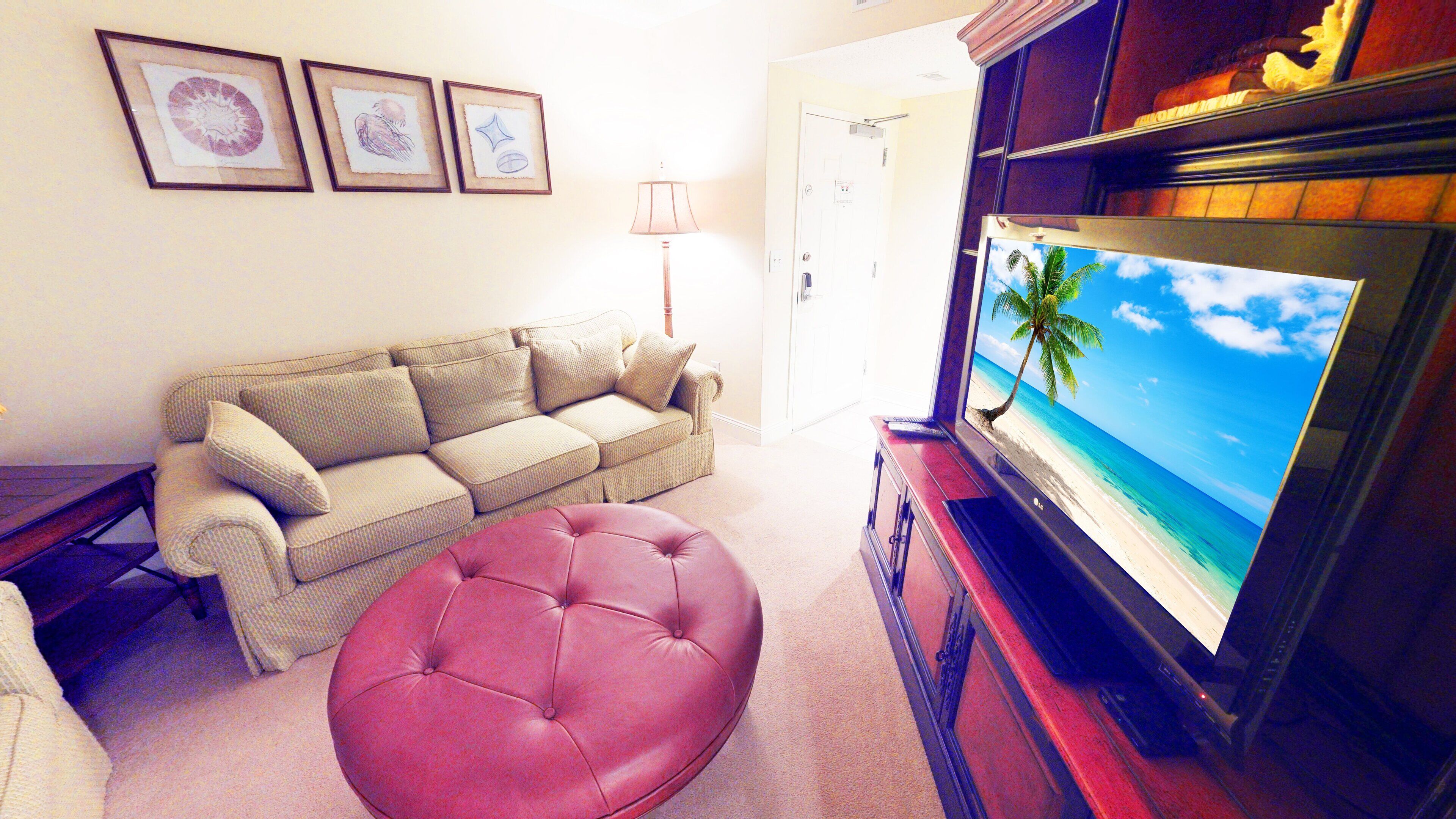 A living room with a beige sofa, a red ottoman, a television displaying a beach scene, and framed artwork on the wall.