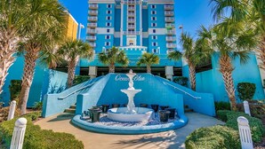 A hotel entrance with a fountain, palm trees, and a blue building with balconies. - Ocean Blue 1101 (Myrtle Beach)