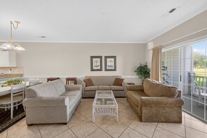A spacious living room with a dining area, a glass coffee table, and a large window with curtains. - Magnolia Place 4673-103 (Myrtle Beach)