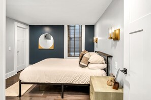 Un dormitorio con una cama, un espejo redondo, una ventana con persianas y una mesita de noche con un reloj.