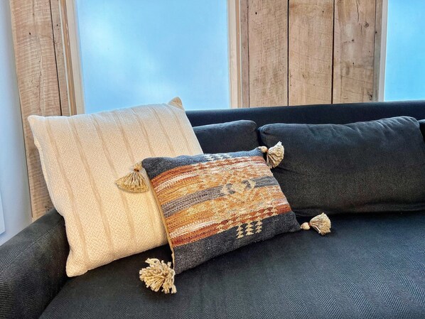 Un sofá oscuro con dos almohadones decorativos, uno beige y otro multicolores con borlas.