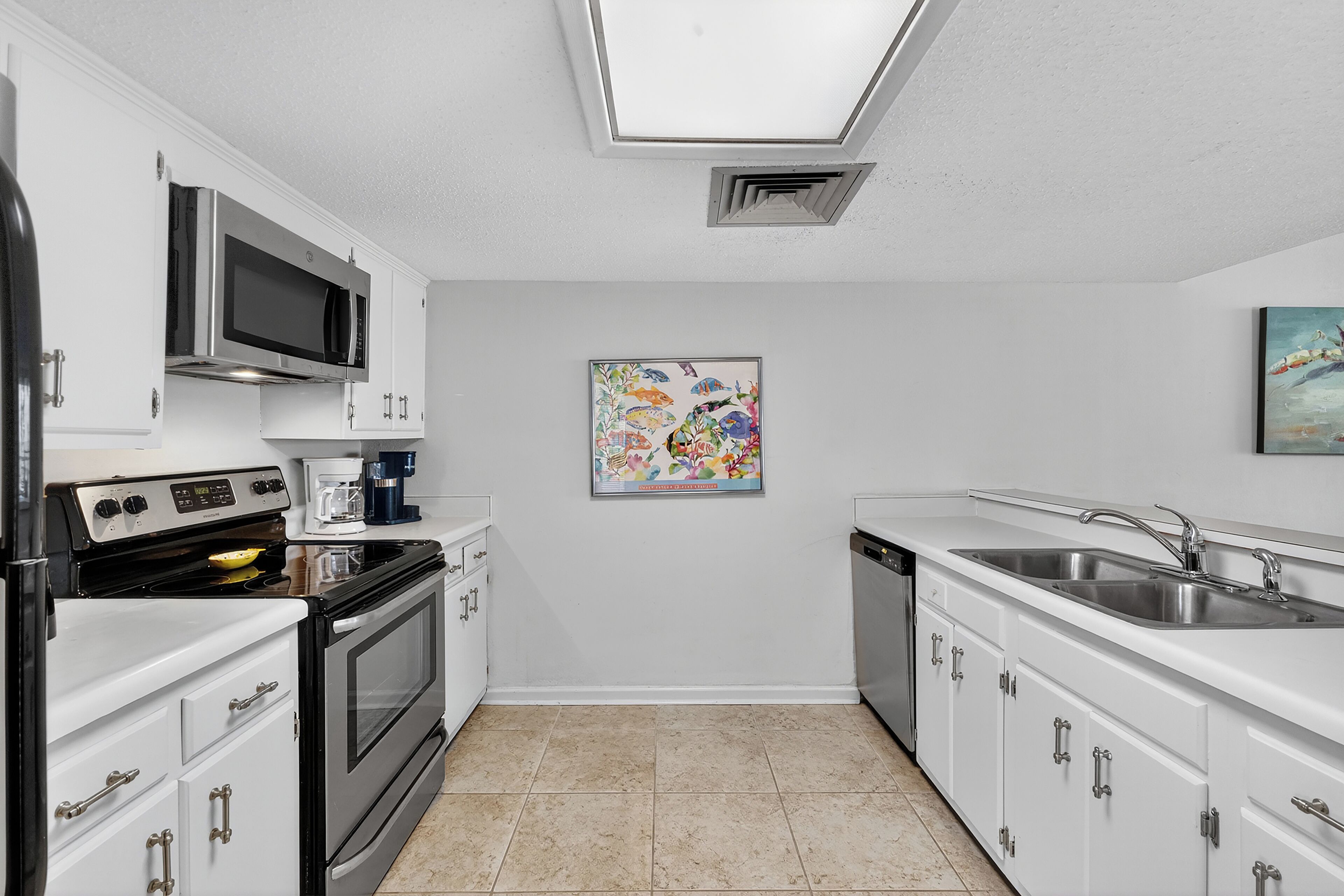 A compact kitchen with white cabinets, a microwave, stove, and a dishwasher. There is a skylight above the sink and a colorful painting on the wall.