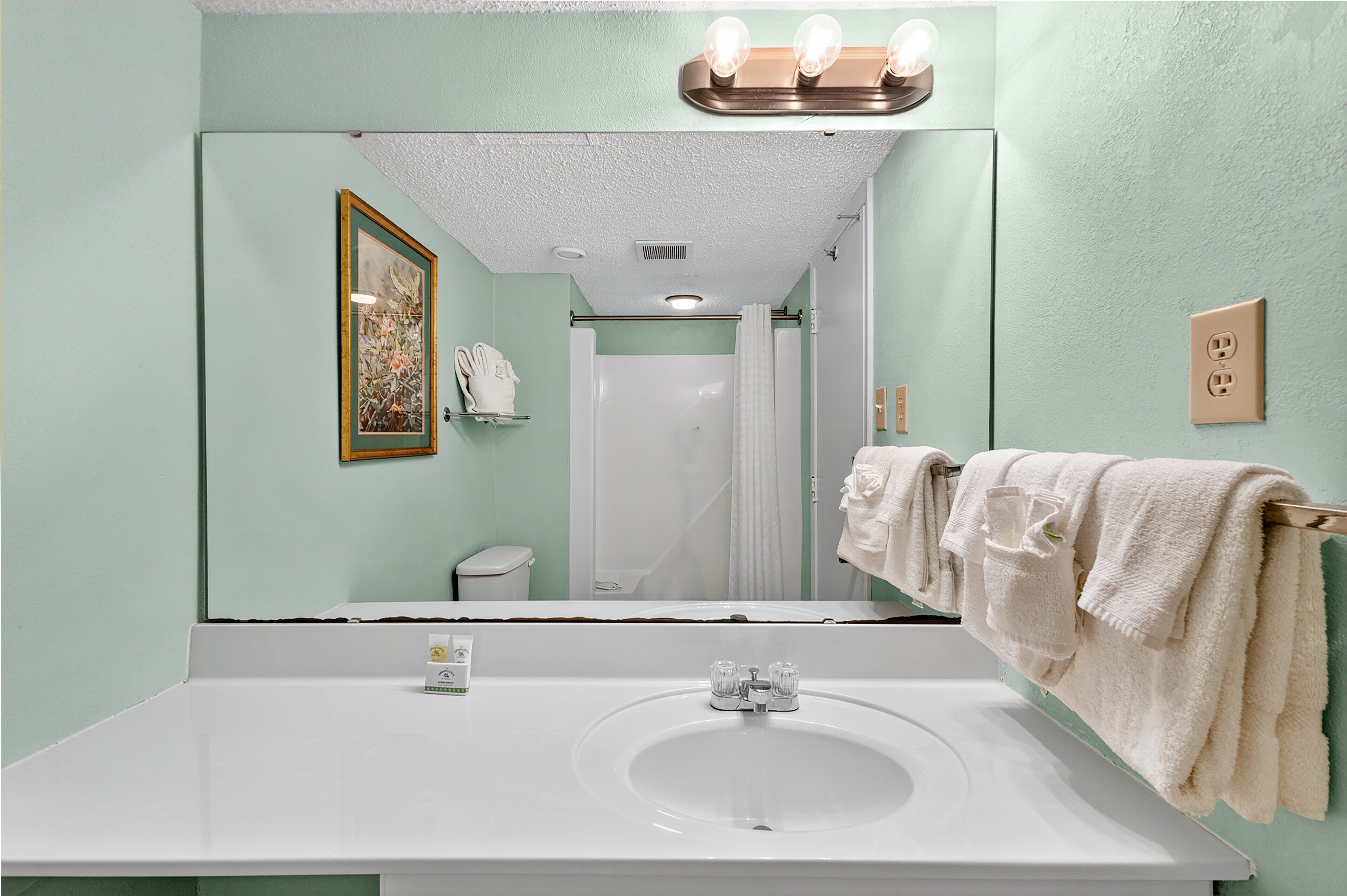 Un baño con lavamanos blanco, un espejo, una lámpara sobre el espejo y un perchero con toallas.