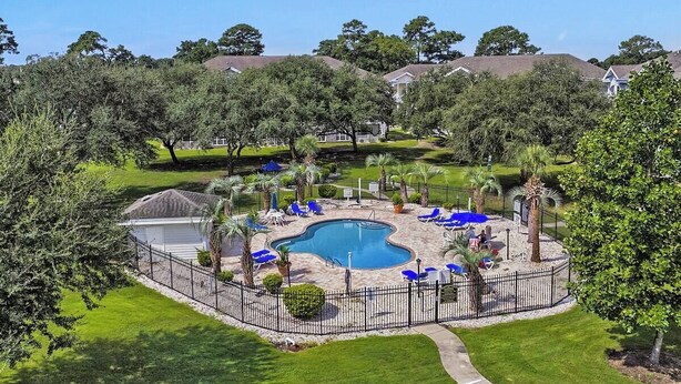 A residential area with a swimming pool surrounded by a fence, palm trees, and houses in the background. - Magnolia Place 4665-104 (Myrtle Beach)