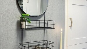 Un baño con tocador de madera, un espejo redondo, estante de alambre negro con una planta en maceta, una vela encendida y una revista sobre el tocador.