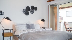 Un dormitorio con una cama de madera, dos mesitas de noche con lámparas, un adorno en la pared y vistas a un balcón exterior.
