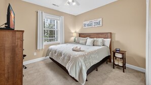 A bedroom with a large bed, a ceiling fan, a window with curtains, a bedside table with a lamp, and a sign on the wall. - Magnolia Place 4691-103 (Myrtle Beach)