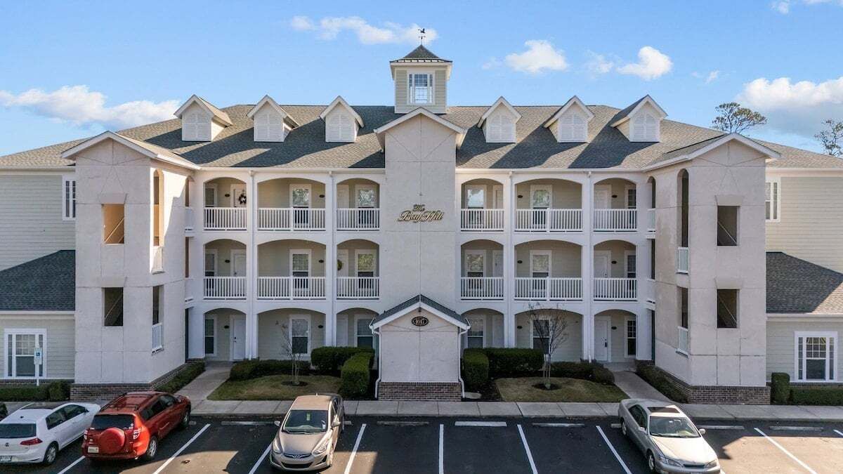 A multi-story hotel with a central entrance, multiple balconies, and a parking lot with several cars.