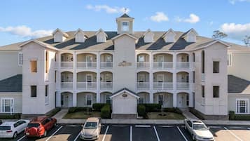 A multi-story hotel with a central entrance, multiple balconies, and a parking lot with several cars.