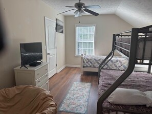 Une chambre avec un lit superposé, un téléviseur posé sur une commode, une fenêtre avec des stores et un ventilateur de plafond.