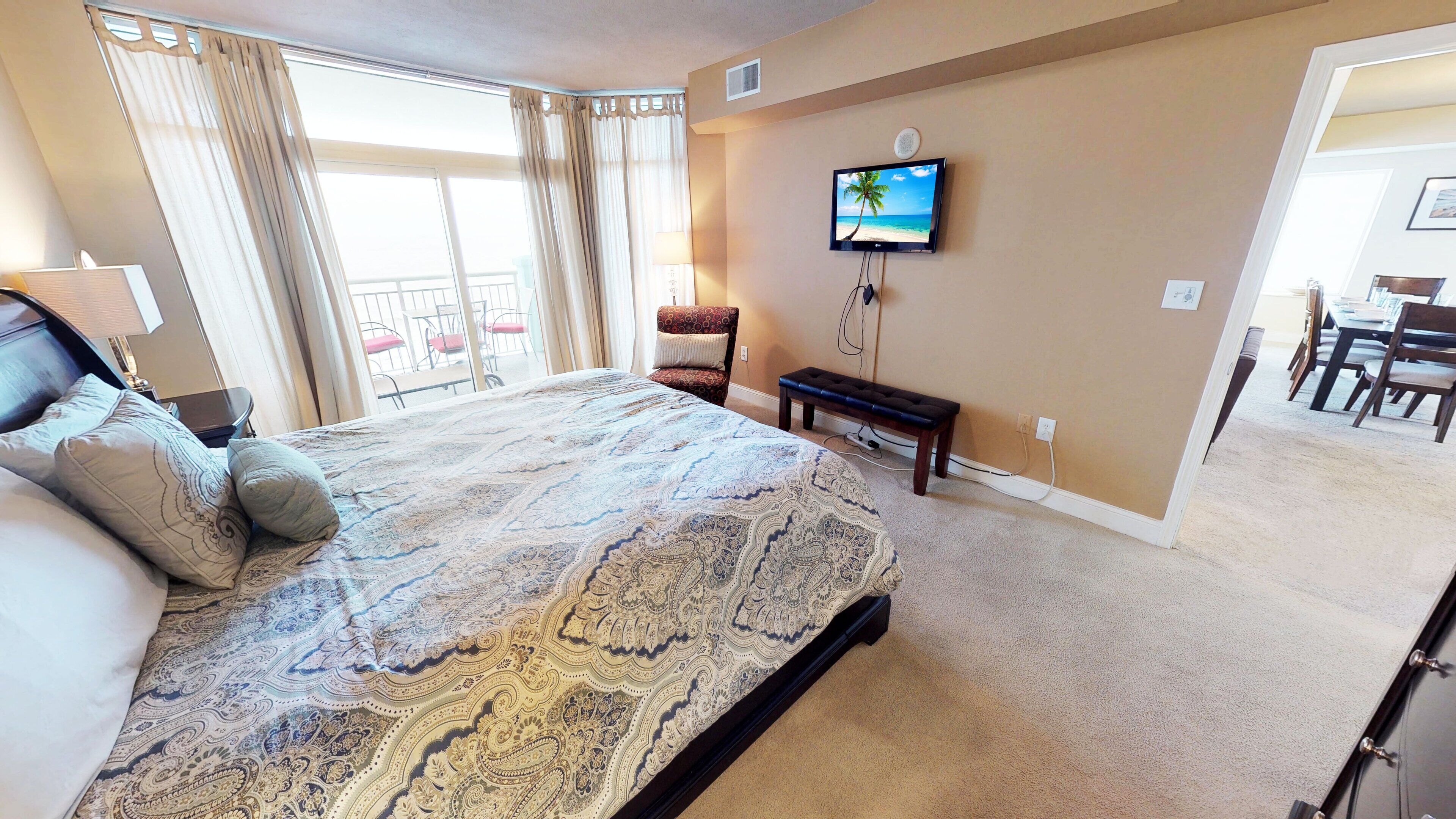 A hotel room with a large bed, a TV mounted on the wall, a dining area with chairs, and a view of the outdoors through the window.