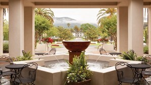 A covered outdoor area with a central water feature, surrounded by tables and chairs, and a view of a distant building and greenery.