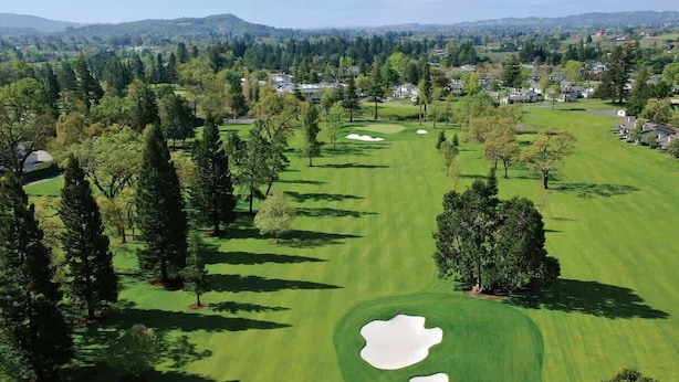 An aerial view of a golf course with sand bunkers, trees, and residential areas in the background.