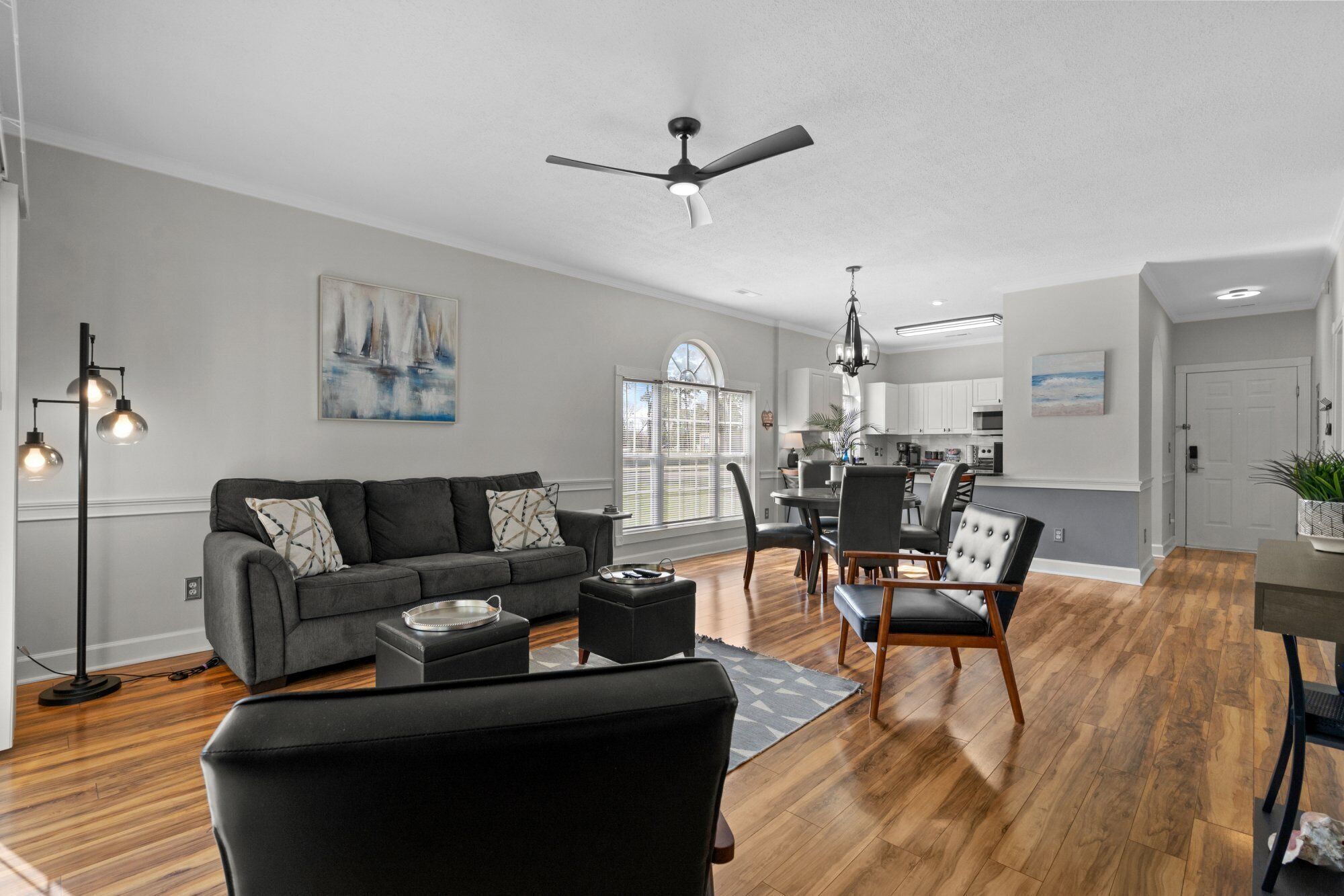 A modern living room with a gray sofa, a dining area with a table and chairs, a ceiling fan, and a view of the kitchen.