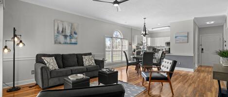 A modern living room with a gray sofa, a dining area with a table and chairs, a ceiling fan, and a view of the kitchen.