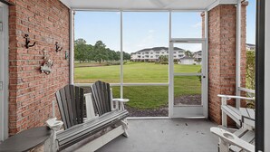 A covered porch with a brick wall, a bench, and a view of a grassy field and buildings in the distance. - Magnolia North 4819-105 (Myrtle Beach)