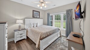 A bedroom with a bed, nightstand, lamp, ceiling fan, TV, and a window with curtains. - Magnolia North 4819-105 (Myrtle Beach)