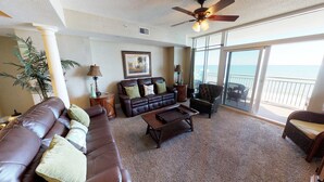 A spacious living room with a leather sofa, a coffee table, and a chair. There is a ceiling fan, a lamp, and a framed picture on the wall. The room has large windows with a view of the ocean. - Ocean Blue 602 (Myrtle Beach)