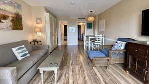 A living room with a sofa, coffee table, and wicker furniture. There is a dining area with a table and chairs. The room has wooden flooring and a TV mounted on the wall. - Ocean Dunes 6011 (Myrtle Beach)