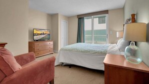 A hotel room with a bed, a television on a wooden dresser, a pink chair, and a lamp on a side table. - Horizon 701 (Myrtle Beach)