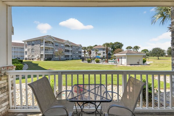 A balcony with two chairs and a table, overlooking a well-maintained lawn and a building with multiple balconies and a small structure. - Magnolia Place 4687-102 (Myrtle Beach)