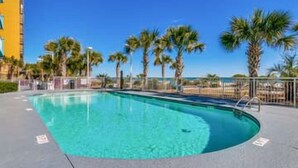 A swimming pool with a clear blue water, surrounded by palm trees and a building in the background. - Ocean Blue 401 (Myrtle Beach)