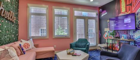 Une pièce avec un mur rose, trois fenêtres équipées de stores, un fauteuil turquoise, un fauteuil bleu, une table basse blanche et un téléviseur fixé au mur.