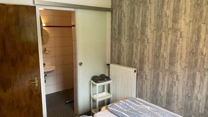 Une chambre à coucher avec un lit, une armoire en bois, un radiateur et une salle de bain visible à travers une porte ouverte.