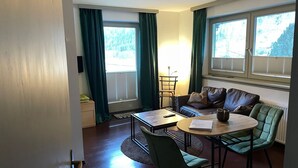 Un salon confortable avec un canapé, une petite table et un fauteuil. Il y a deux fenêtres avec des rideaux, une lampe et un radiateur.