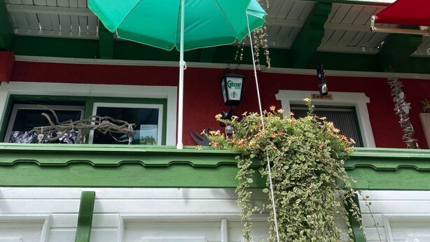 Un immeuble à plusieurs étages aux façades vertes et blanches, doté de balcons ornés de plantes suspendues et surmonté d’un parasol vert.