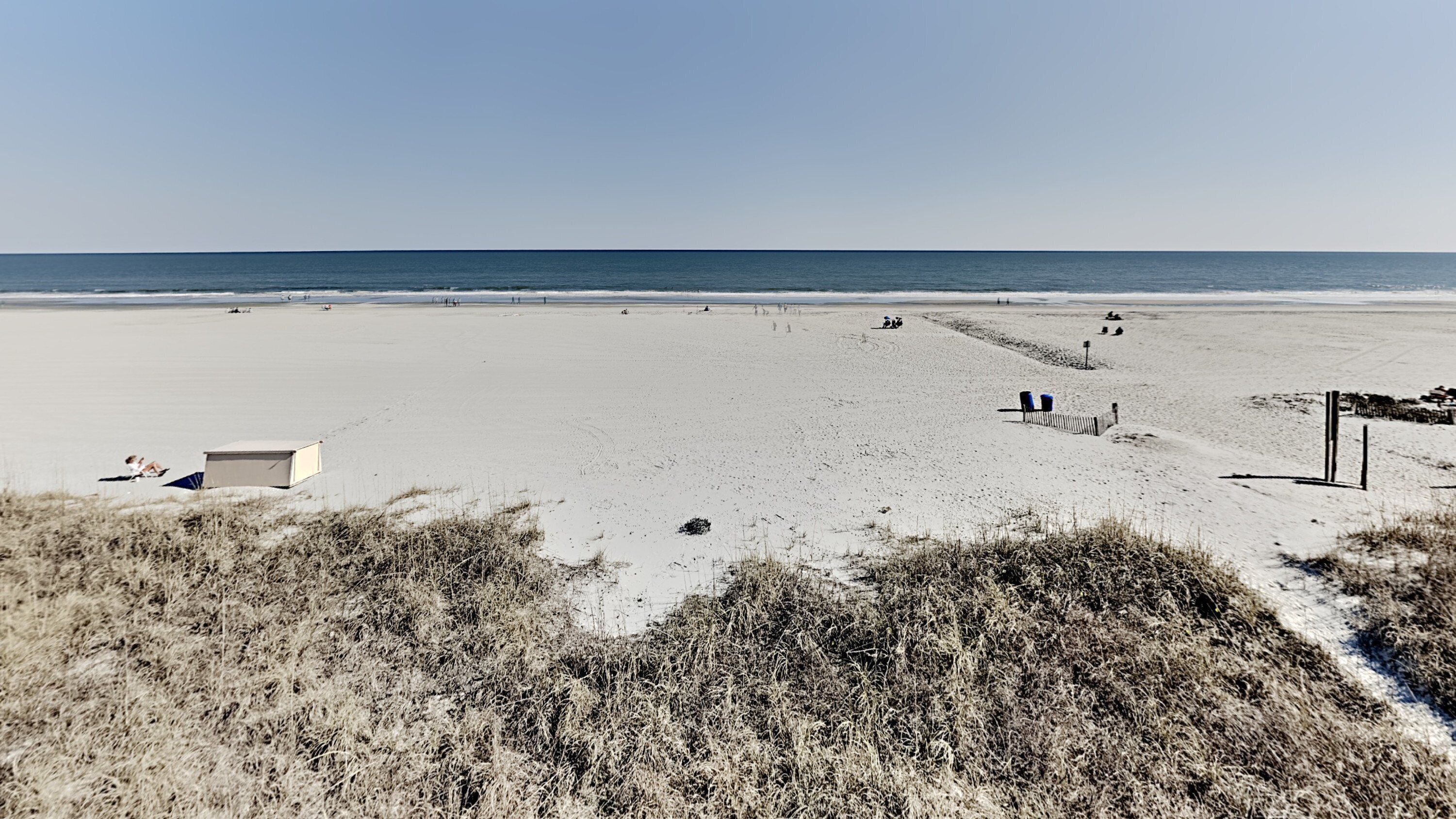 A wide sandy beach with a few people scattered across it, a small building, and a clear sky.