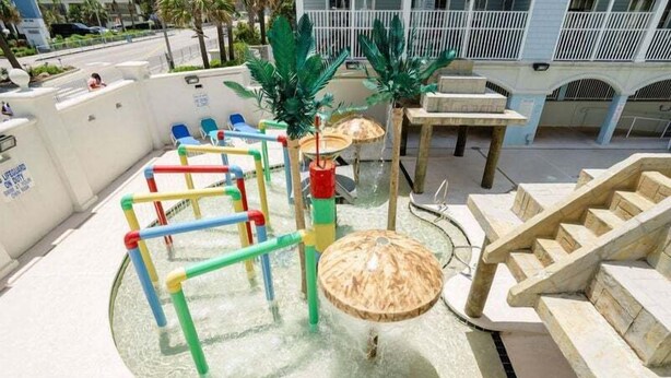 Un espace de jeux aquatiques avec des toboggans et des structures d’escalade, entouré d’un mur blanc et agrémenté de plantes en pot.