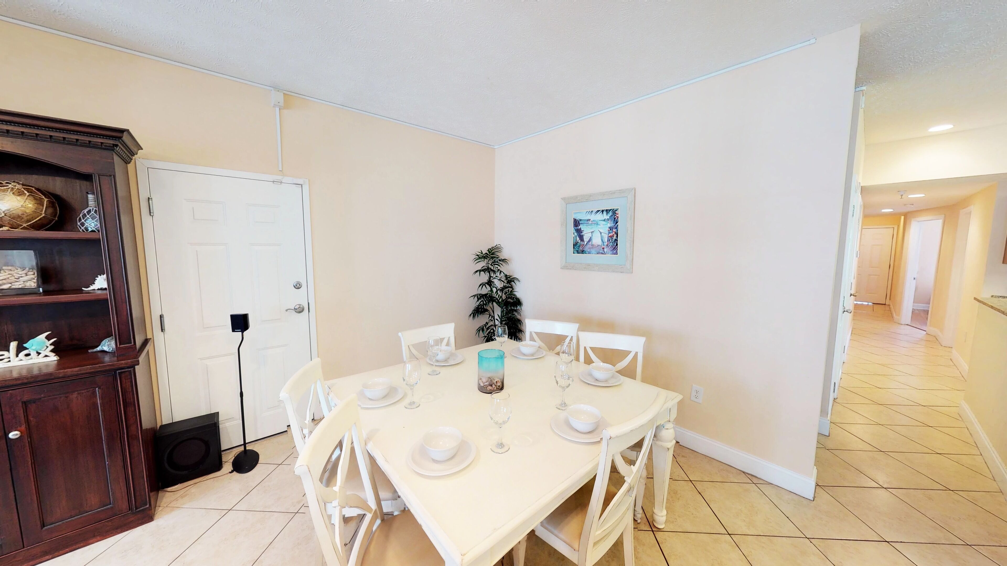 A dining area with a white table set for four, surrounded by white chairs. There is a wooden cabinet with decorative items on the left. A potted plant is in the center of the room.