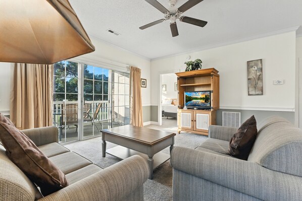 A spacious living room with a ceiling fan, a television on a wooden stand, and a sliding glass door leading to an outdoor area. - Magnolia Place 4647-201 (Myrtle Beach)