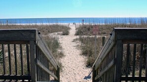 Un sentier menant à la plage et à l’océan, bordé de rambardes en bois, et sur lequel est apposée une pancarte indiquant : « Parasols uniquement ».