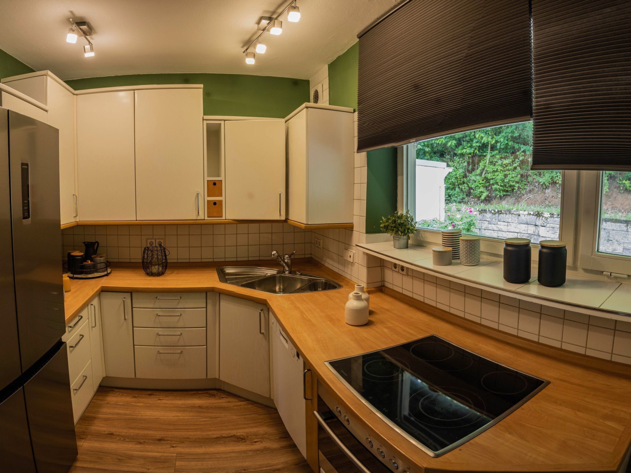 Eine moderne Küche mit weißen Schränken, einem Edelstahlkühlschrank, einem eingebauten Backofen und einem Fenster mit Blick auf Grünflächen.