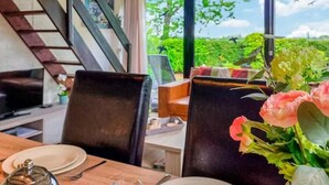 Uma sala de jantar com mesa posta para dois, um vaso de flores e vista para o verde lá fora.
