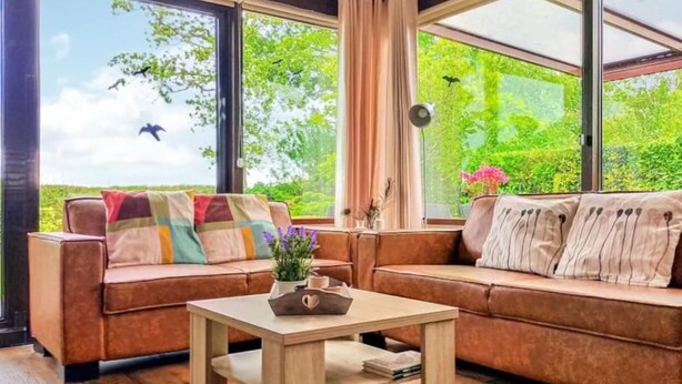 Uma sala de estar aconchegante com um sofá de couro marrom, uma mesa de centro de madeira e janelas amplas com vista para vegetação e pássaros.