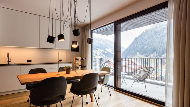 A modern kitchen with a wooden table and chairs, a large window with a mountain view, and a balcony with chairs. - Stillvolle Luxus Wohnung in der Residenz Silvretta by Interhome (See)
