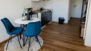 Een compacte keuken met een magnetron, spoelbak en vaatwasser. Twee blauwe stoelen staan rond een kleine tafel met een plant en een fles. Een prullenbak bevindt zich naast de spoelbak.