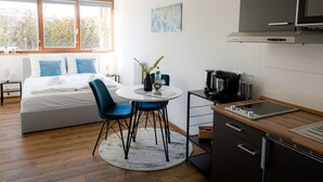 Een moderne slaapkamer met een bed, een kleine tafel met stoelen, een raam en een kitchenette met een magnetron en koffiezetapparaat.