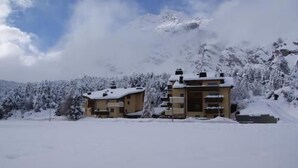 Un chalet à deux étages, dont le toit est recouvert de neige, entouré d’un paysage enneigé et de montagnes.