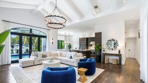 A spacious living room with a large sectional sofa, blue accent chairs, and a round coffee table. A modern chandelier hangs from the ceiling, and a large mirror is mounted on the wall. - The Rendez-Vous Villa (Fort Lauderdale)