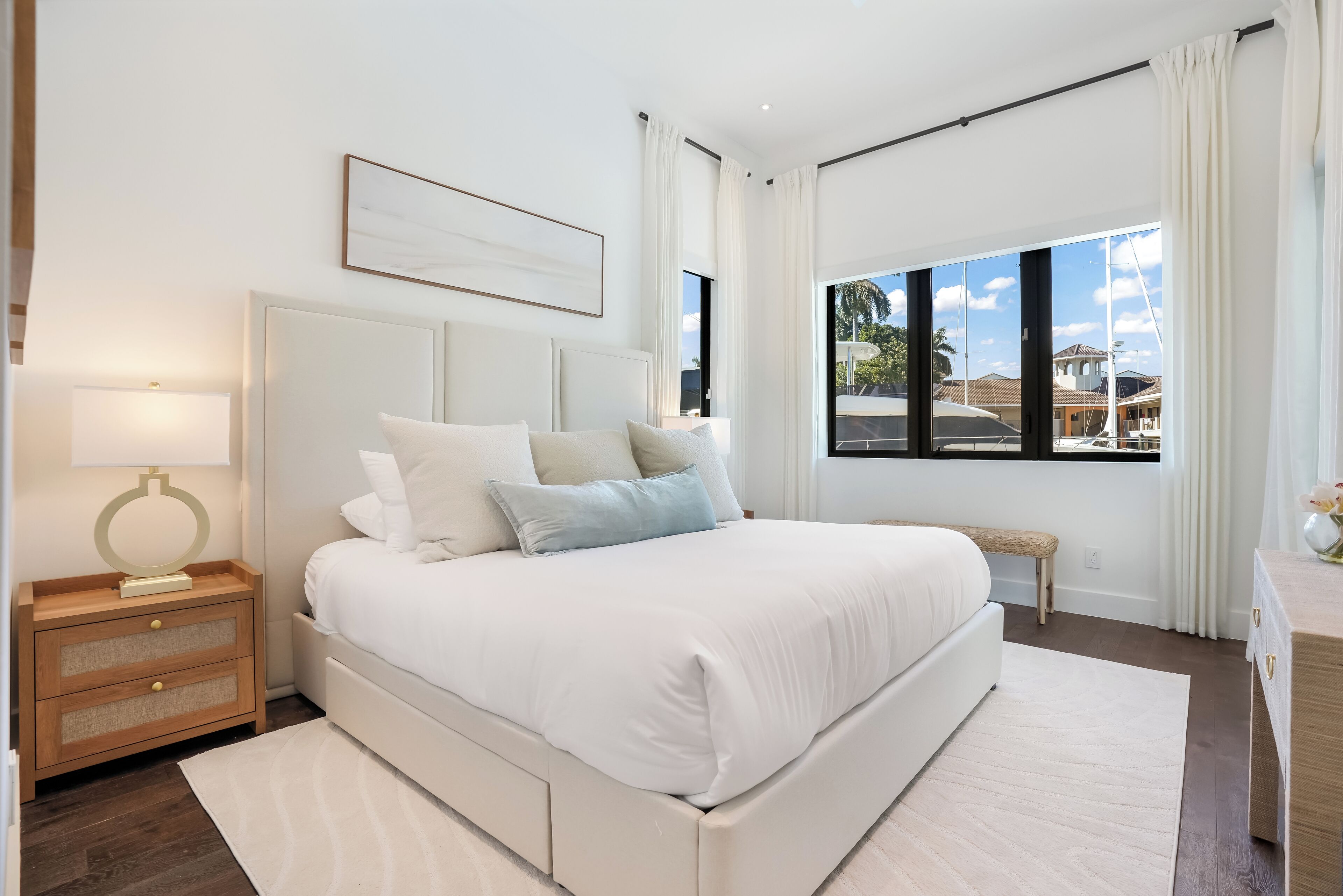 A modern bedroom with a large bed, a bedside table with a lamp, and a view of the sea through large windows.