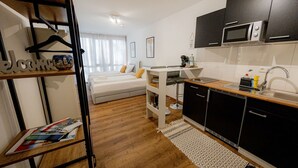 Quarto de hotel pequeno com cozinha compacta, uma cama com almofadas, uma mesinha e uma estante com uma placa de 'Bem-vindo'.